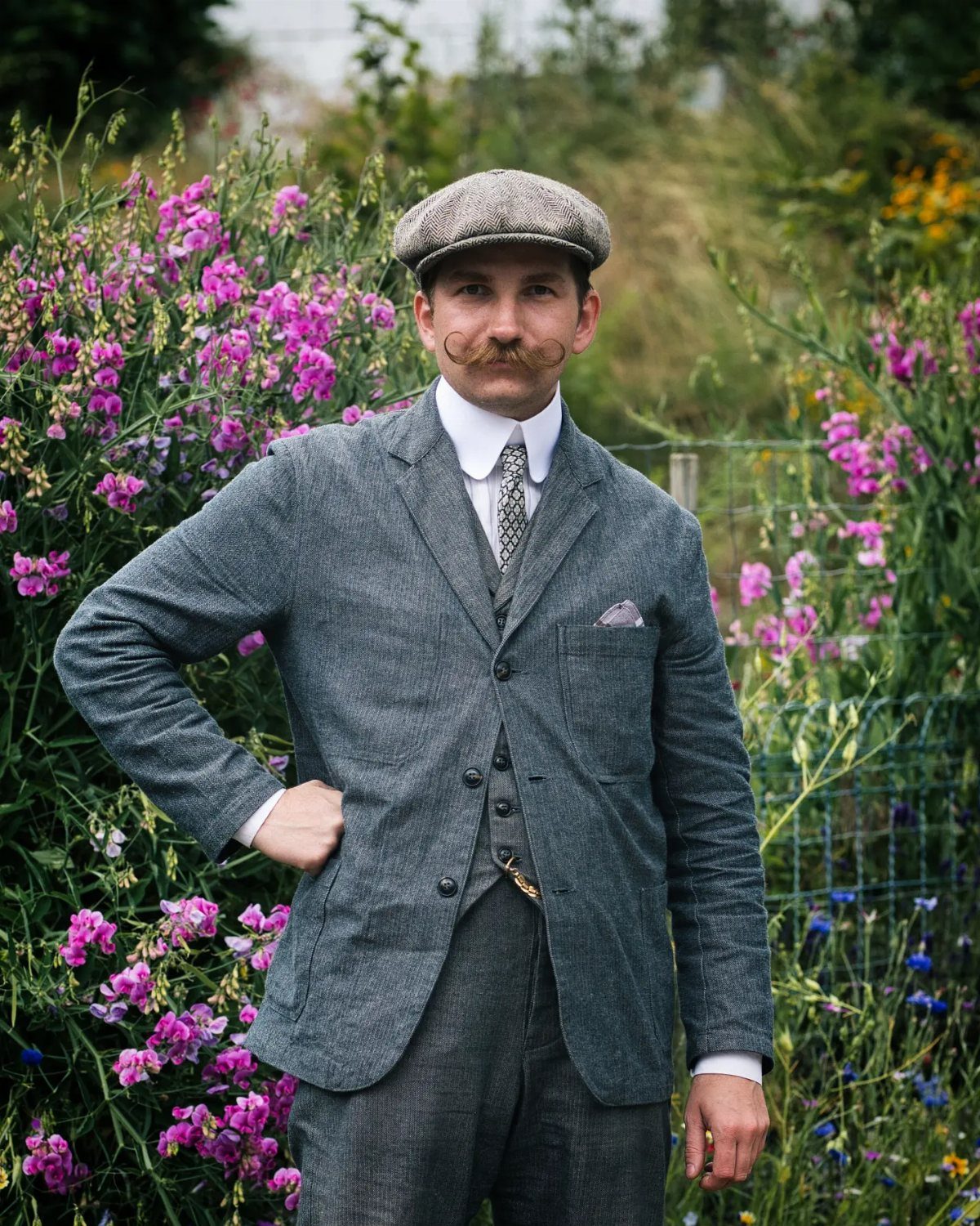 Man in grey tweed three-piece suit and flat cap standing among purple flowers.