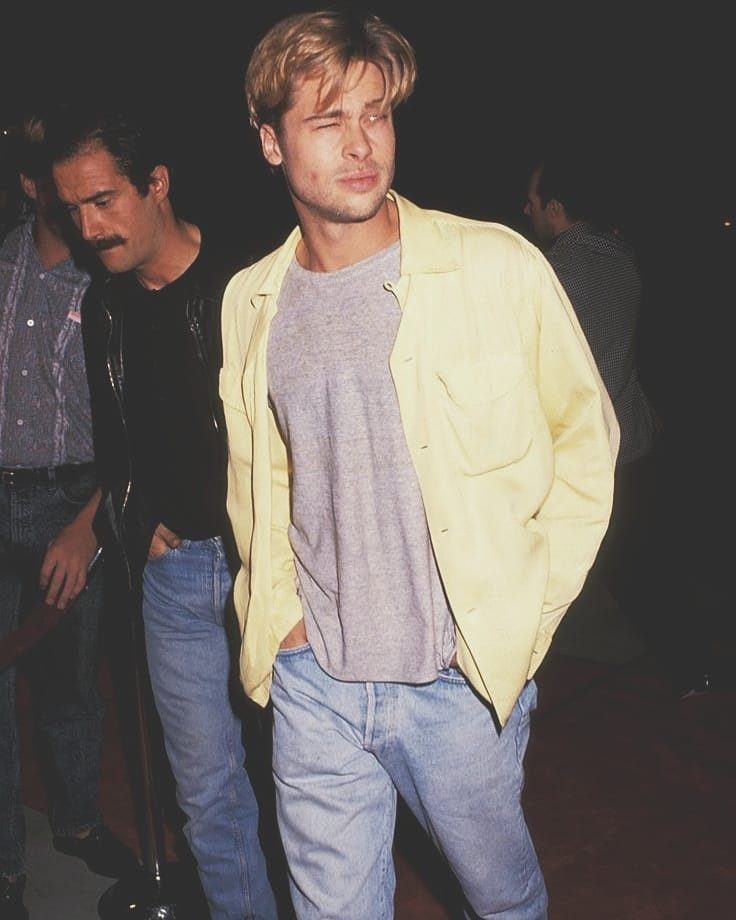 Man in oversized yellow shirt and light blue jeans posing in a dark setting.