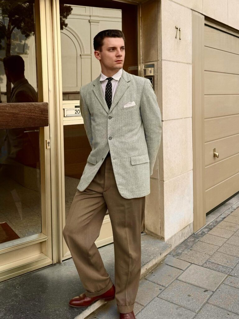Man wearing a 1960s style grey blazer, narrow tie, and brown pleated trousers.