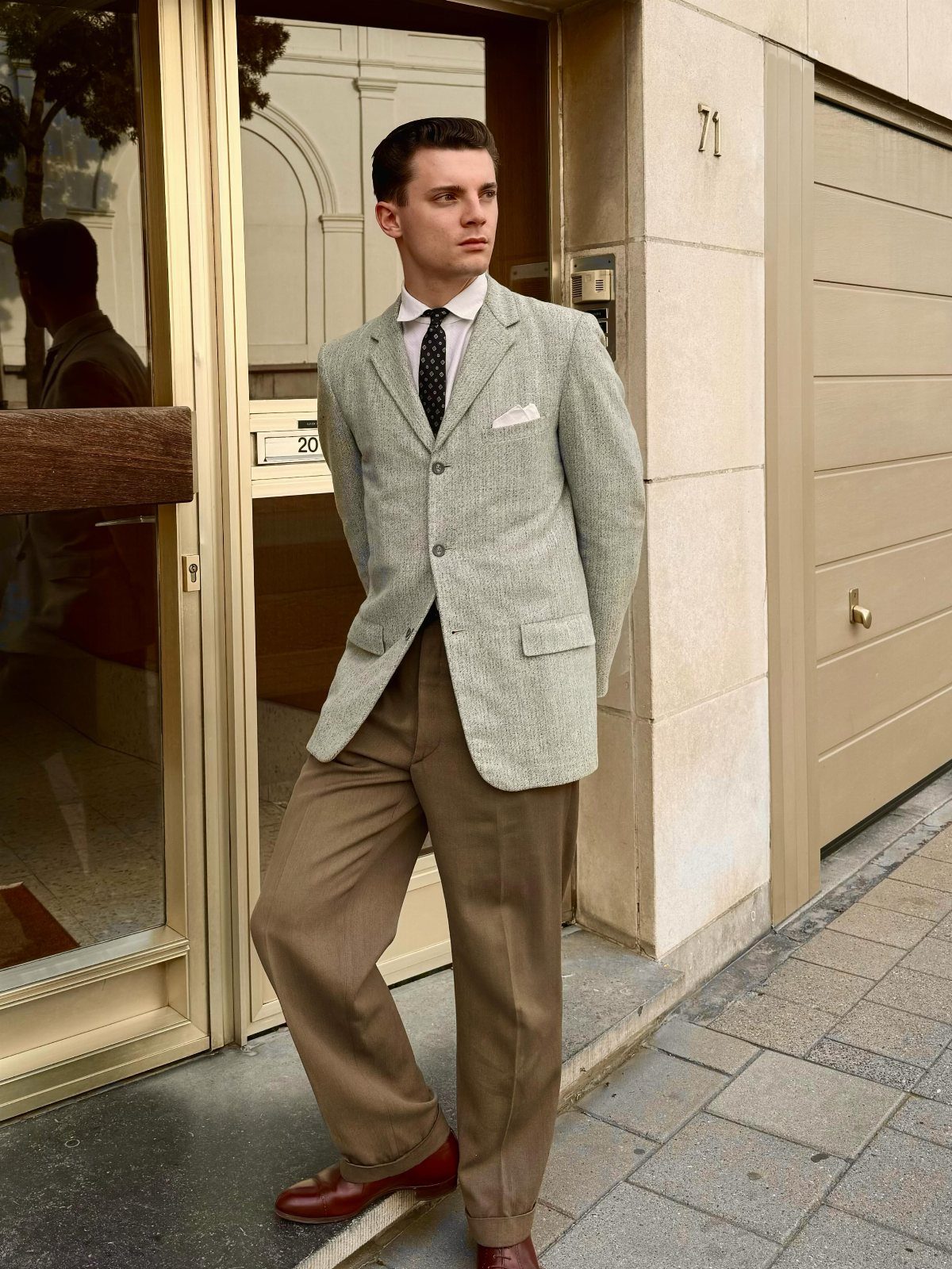 Man wearing a 1960s style grey blazer, narrow tie, and brown pleated trousers.