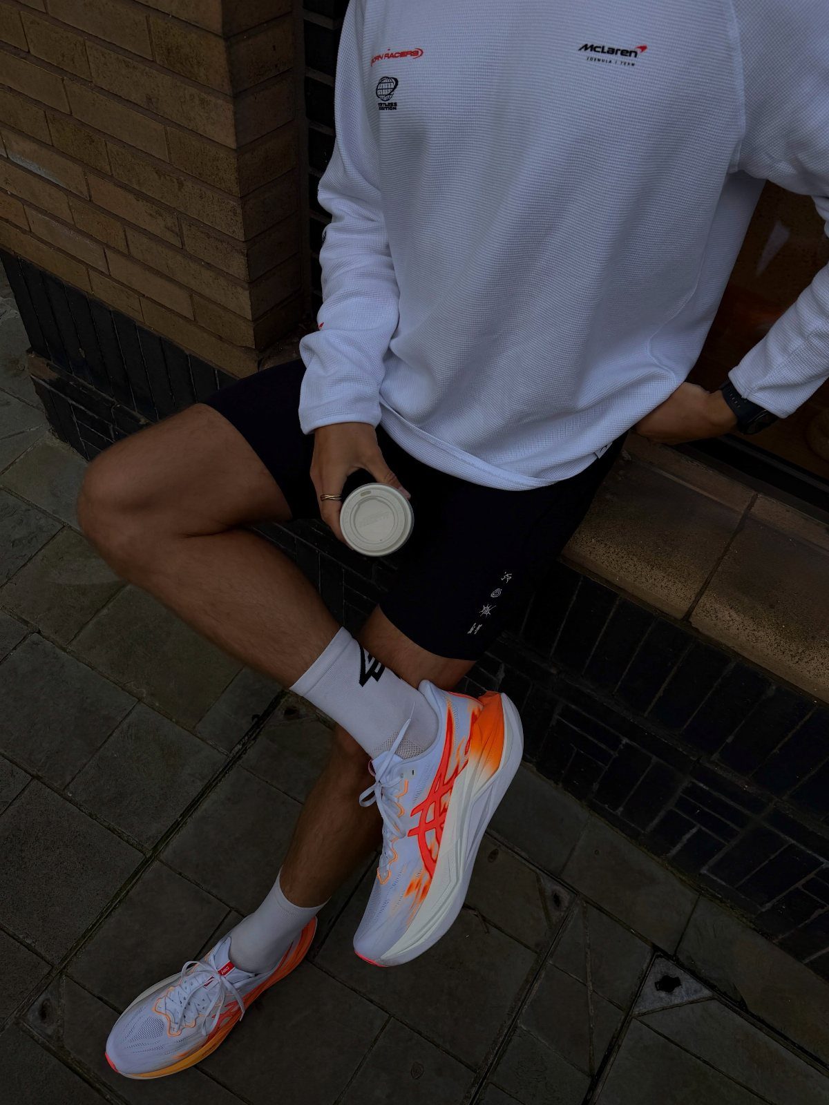 Man in white athletic shirt, black shorts, and orange-accented sneakers sitting outdoors.