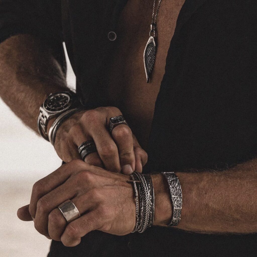 Man wearing silver rings, bracelets, a watch, and a wing pendant necklace.