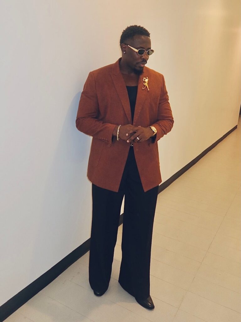 Black man in a rust-colored blazer and wide-leg black trousers posing against a white wall.