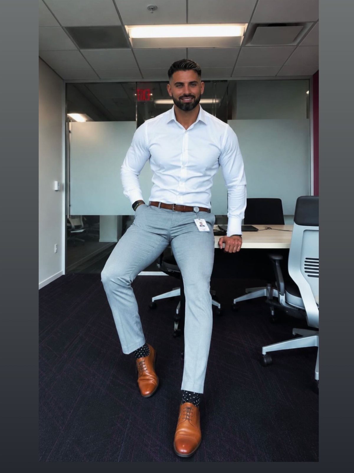 Man in white shirt and grey trousers posing in a modern office setting.