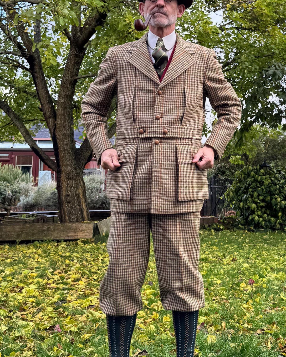 Man in brown checkered Norfolk jacket and knickerbockers smoking a pipe outdoors.