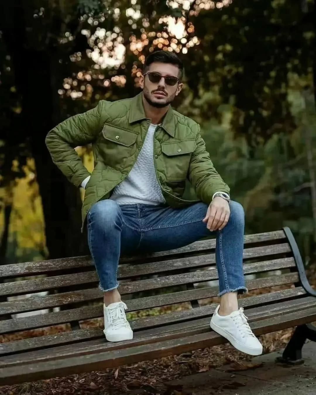 Man in olive quilted jacket, grey sweater, and blue jeans sitting on park bench