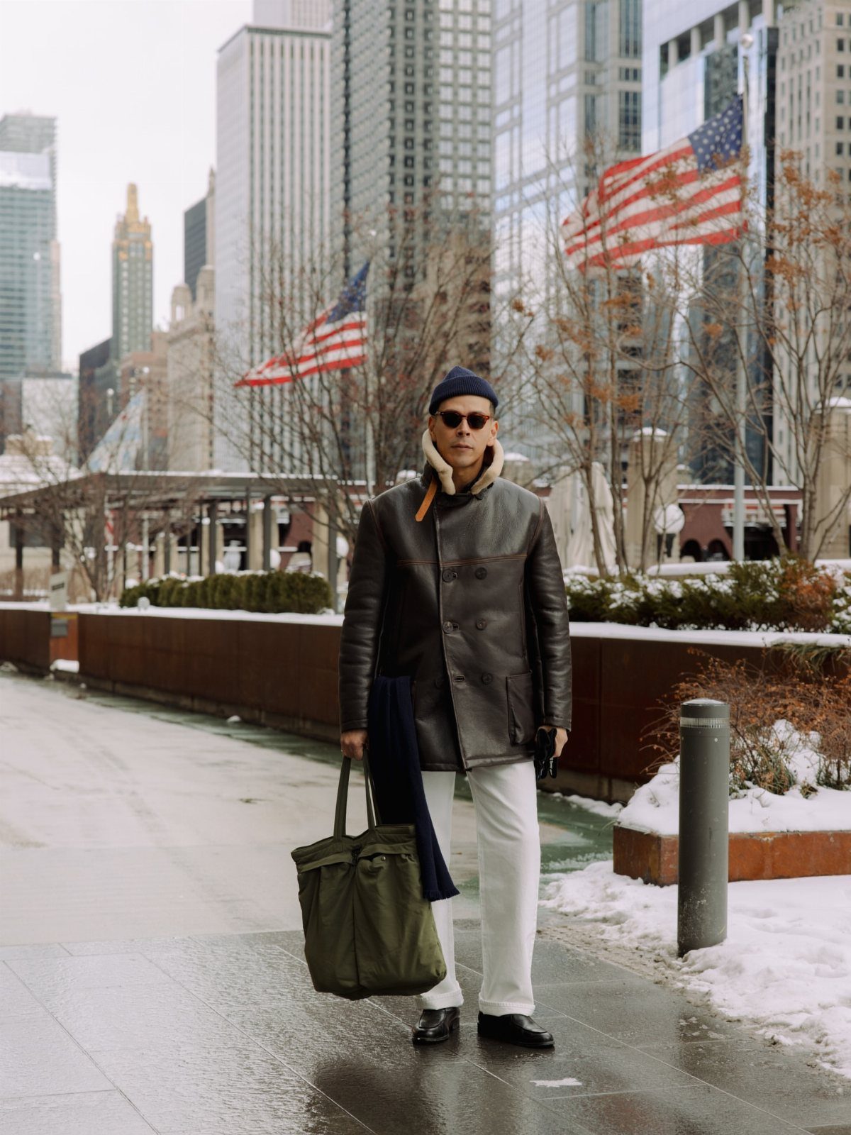 Man wearing leather jacket, scarf, and beanie standing in snowy Chicago city center.