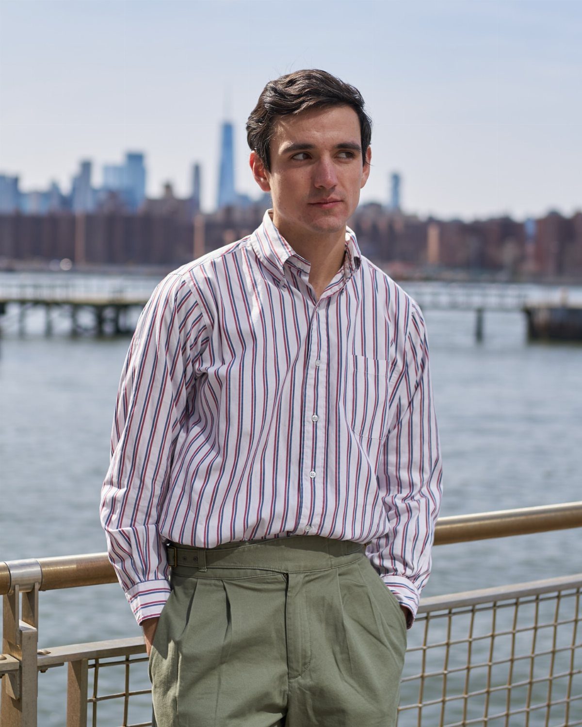 Man wearing a striped oxford shirt and olive green trousers by the water.