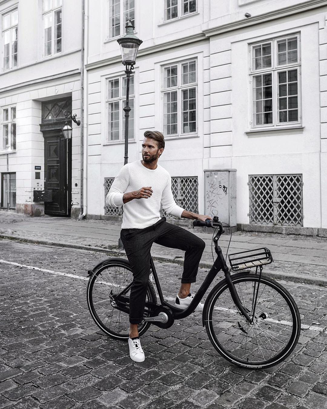 Man in white sweater and black trousers sitting on a bicycle in Copenhagen.