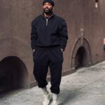 Man in black streetwear, beanie, and chunky beige sneakers posing on a city sidewalk.