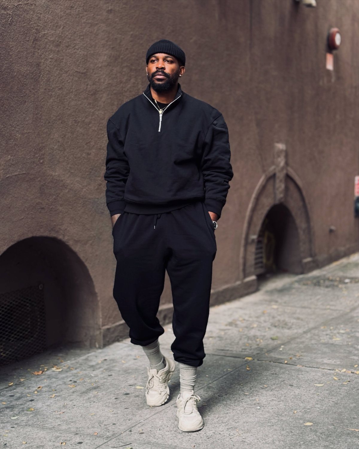 Man in black streetwear, beanie, and chunky beige sneakers posing on a city sidewalk.