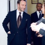 Tom Ford wearing a sleek black suit and brown tie in an indoor setting.