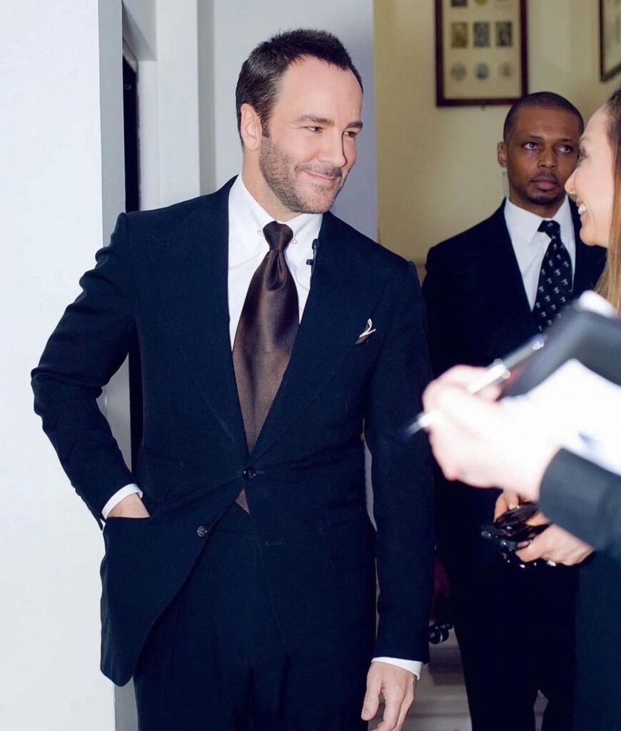 Tom Ford wearing a sleek black suit and brown tie in an indoor setting.