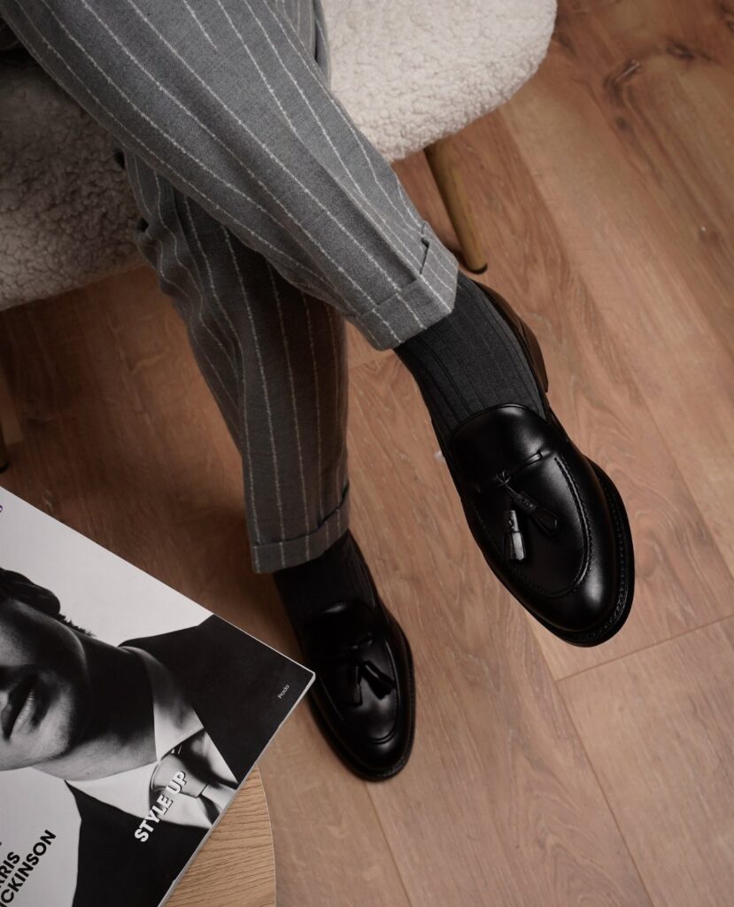 Black leather tassel loafers worn with grey pinstriped trousers and black socks.