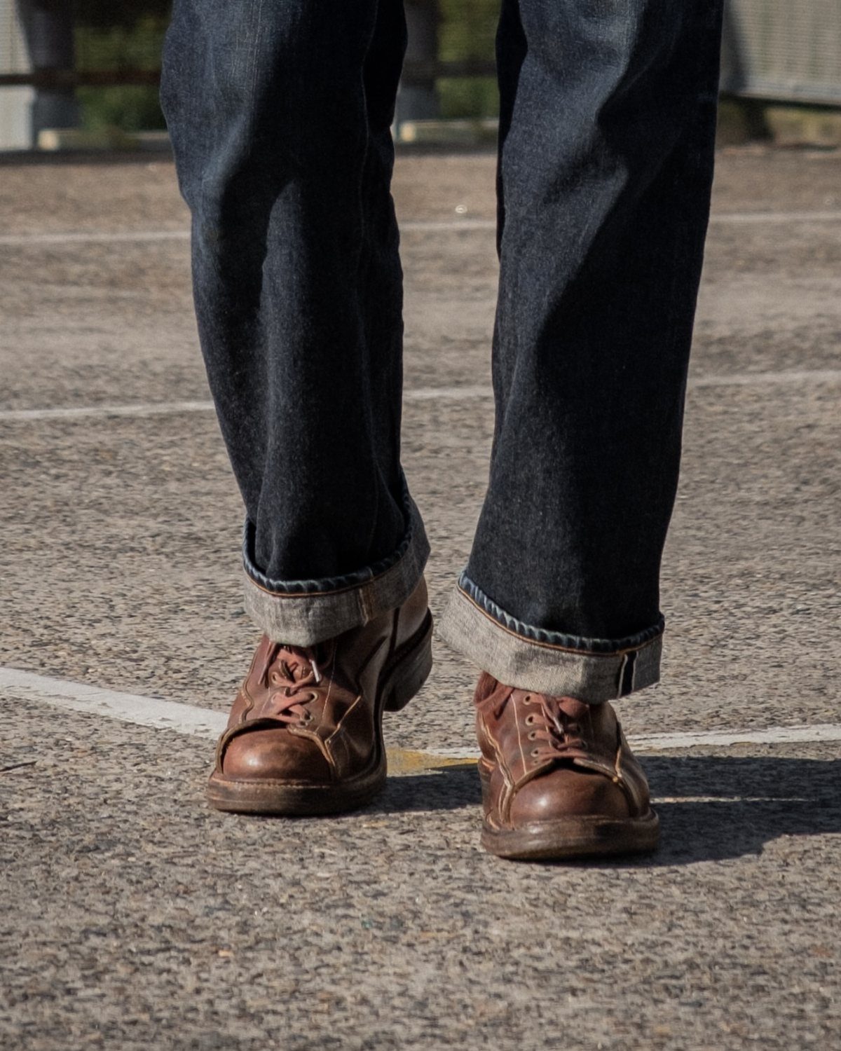 Brown leather boots and cuffed dark blue jeans on a grey asphalt ground.
