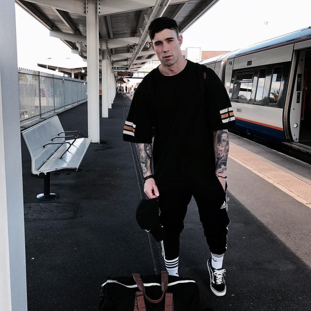 Man in black oversized t-shirt and striped joggers standing on a train platform.