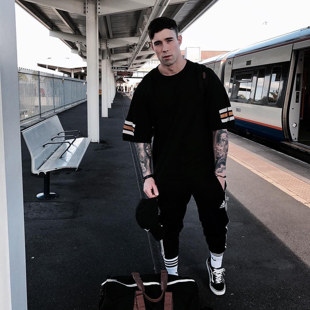 Man in black oversized t-shirt and striped joggers standing on a train platform.