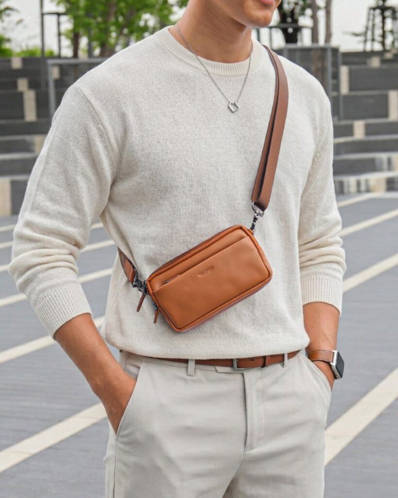 Man wearing a brown leather crossbody bag over a cream sweater and grey trousers.