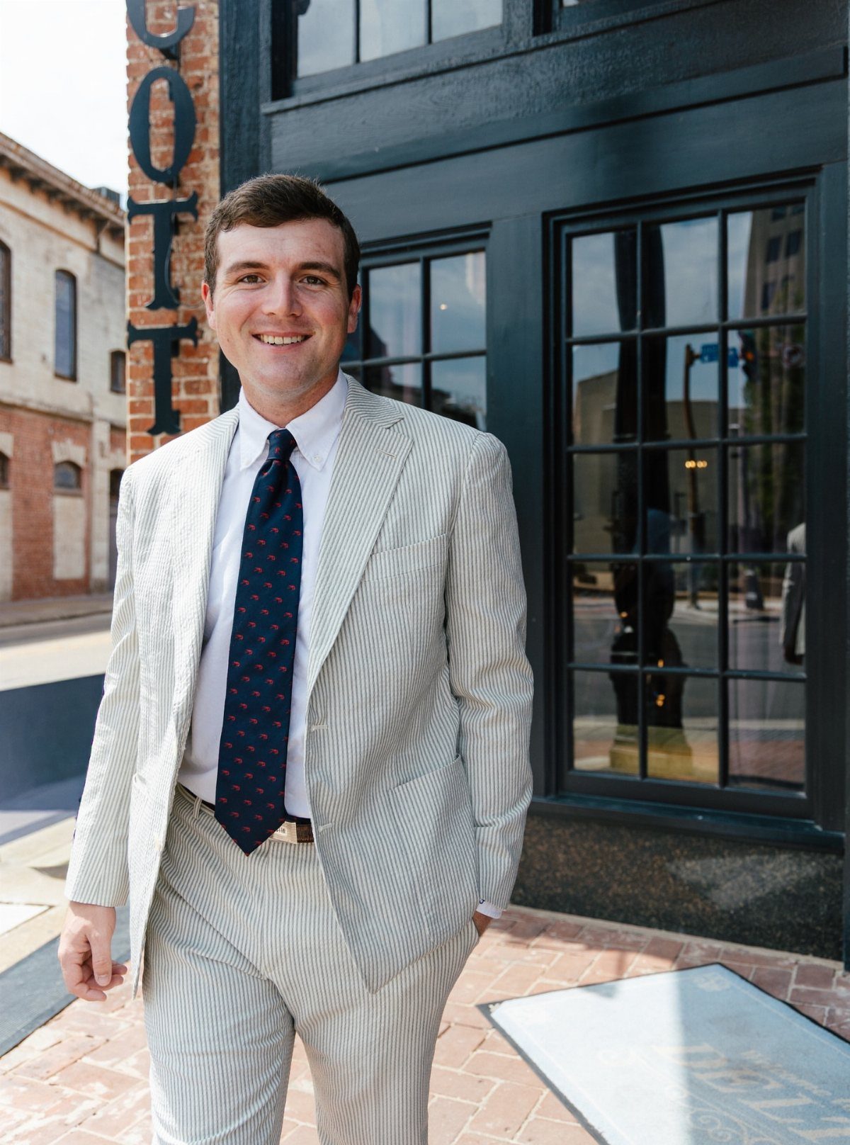 Man in a light pinstriped suit and navy tie smiling in front of a building.