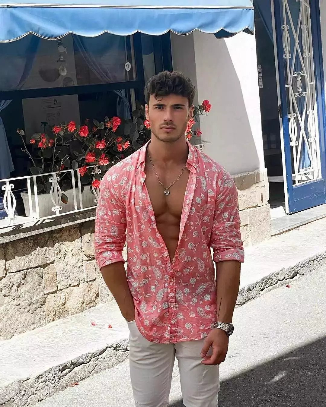 Man in open pink patterned shirt and white pants posing on a sunny street.