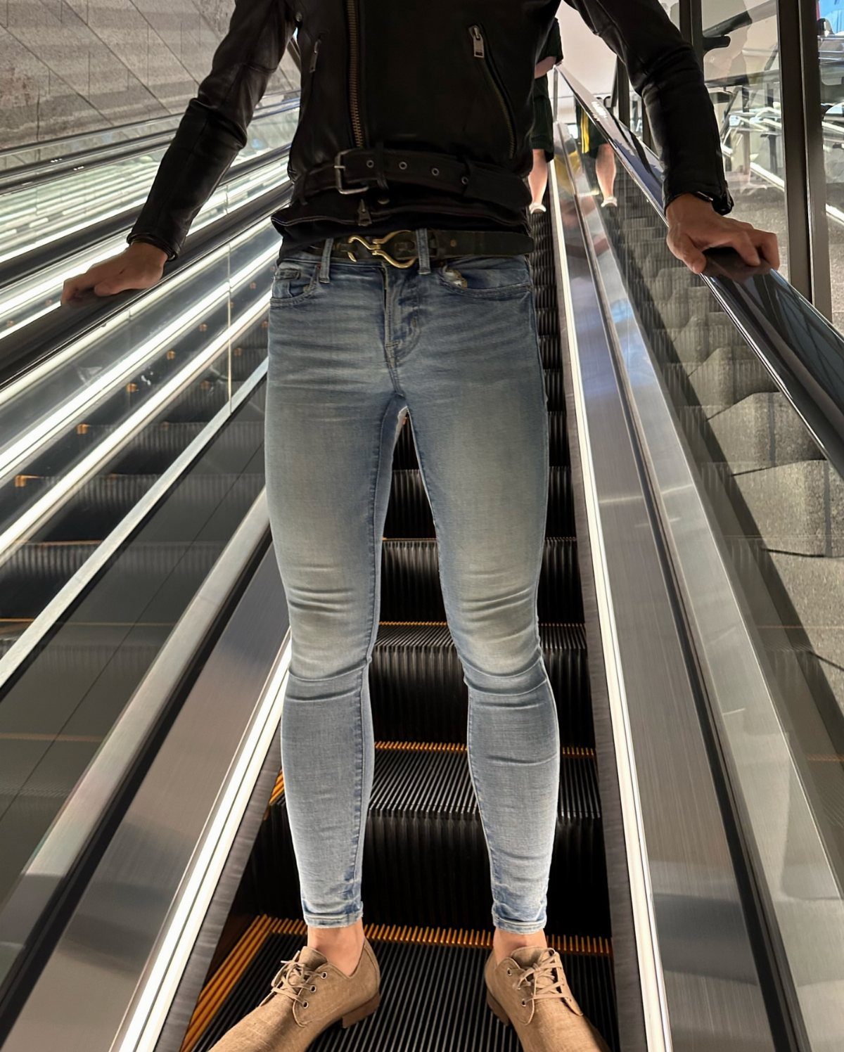 Slender person in leather jacket and skinny blue jeans standing on an escalator.