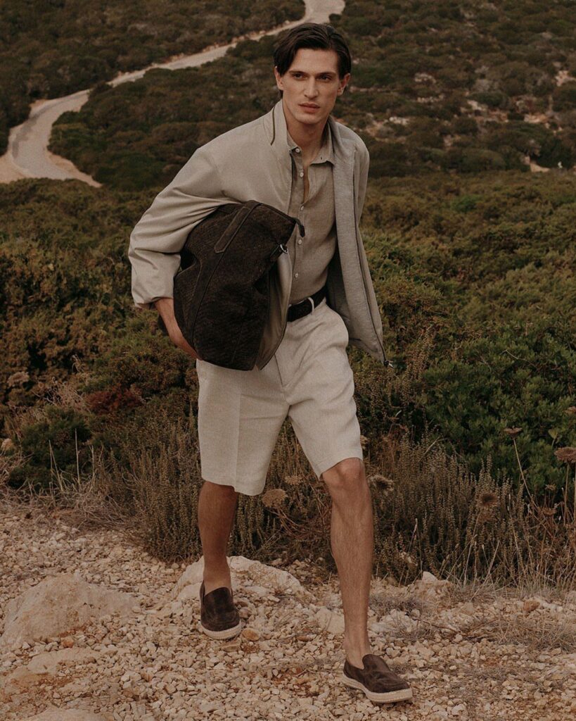 Man in beige shirt and white shorts holding a leather bag on a hillside