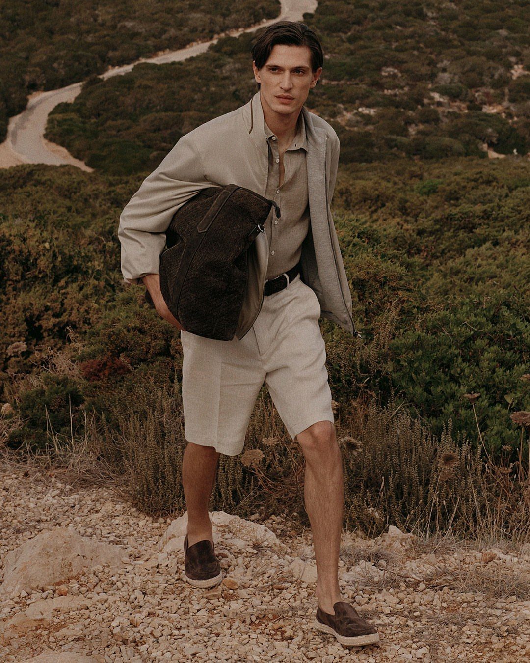 Man in beige shirt and white shorts holding a leather bag on a hillside
