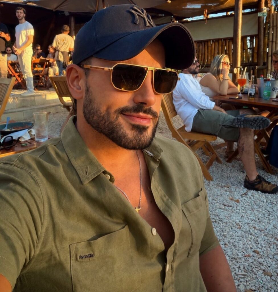Man wearing a dark blue baseball cap and gold sunglasses at an outdoor cafe.