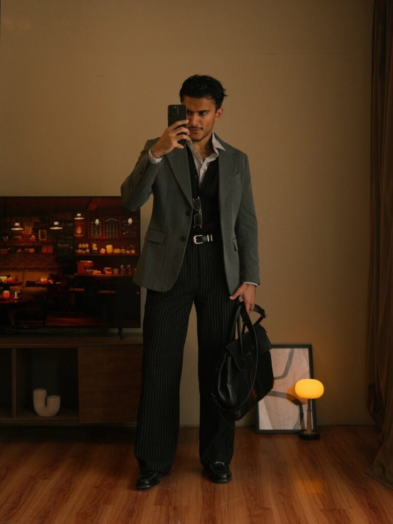 Man in a grey blazer and pinstriped trousers taking a mirror selfie indoors.
