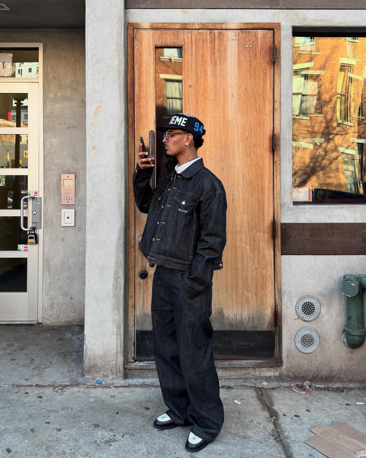 Man in black denim jacket and trousers wearing a baseball cap on city sidewalk