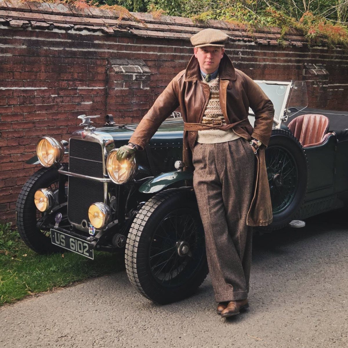 Man in 1920s attire with leather jacket and flat cap leaning on vintage car.