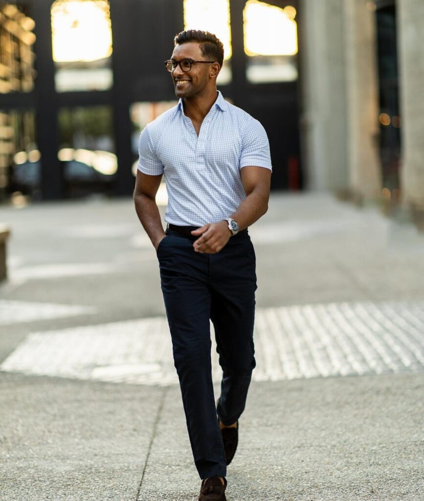 Man in a blue checkered short-sleeve shirt and navy trousers walking on a city sidewalk.