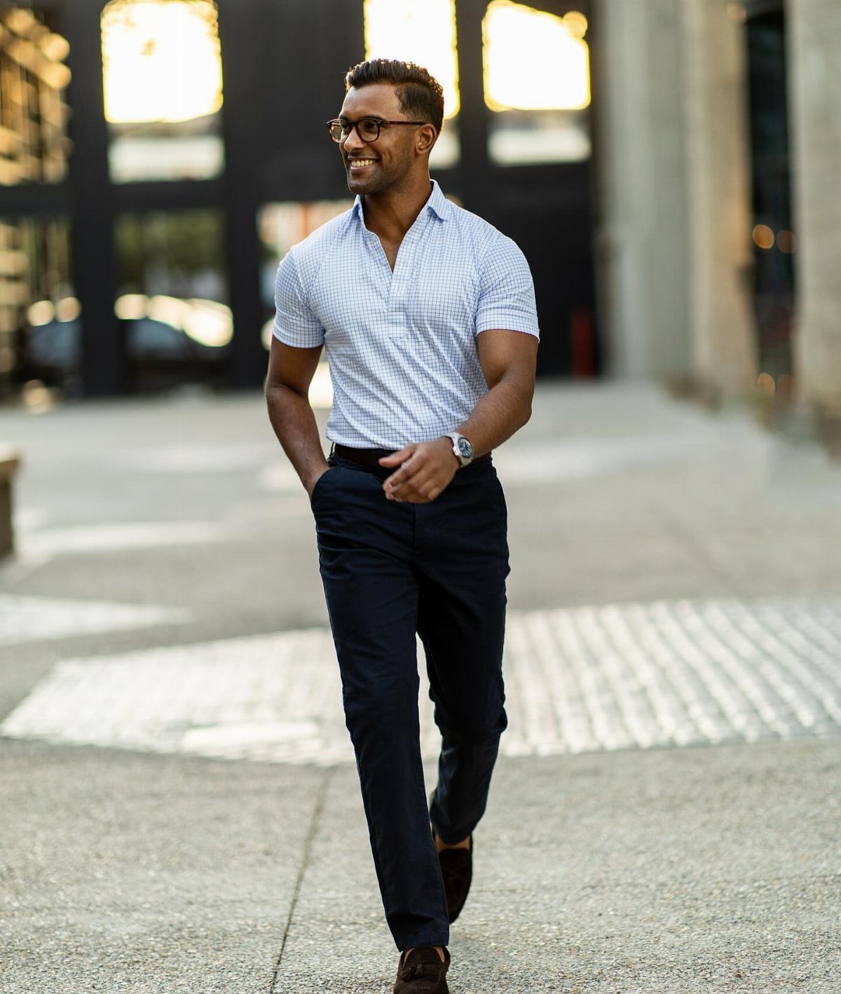 Man in a blue checkered short-sleeve shirt and navy trousers walking on a city sidewalk.
