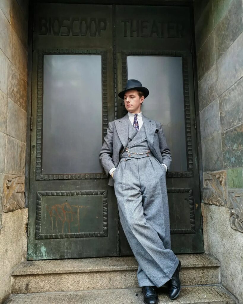 Man in a grey vintage three-piece suit and fedora leaning against theater doors.
