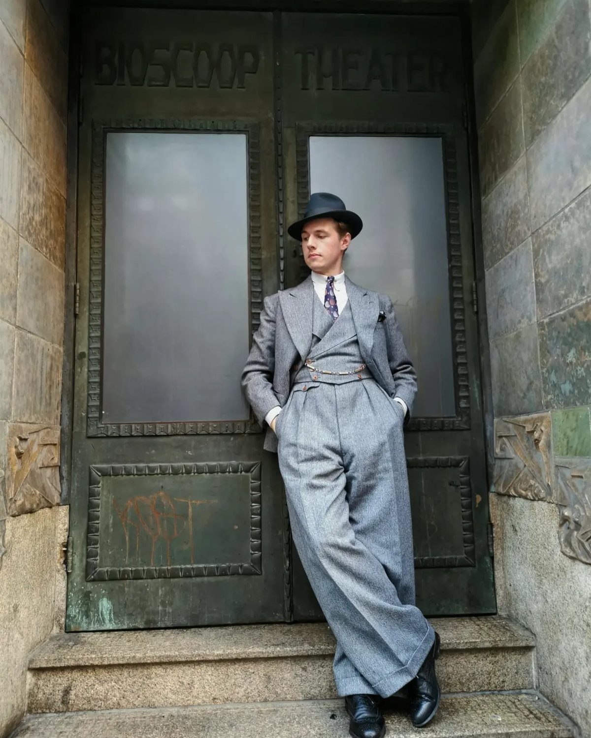 Man in a grey vintage three-piece suit and fedora leaning against theater doors.